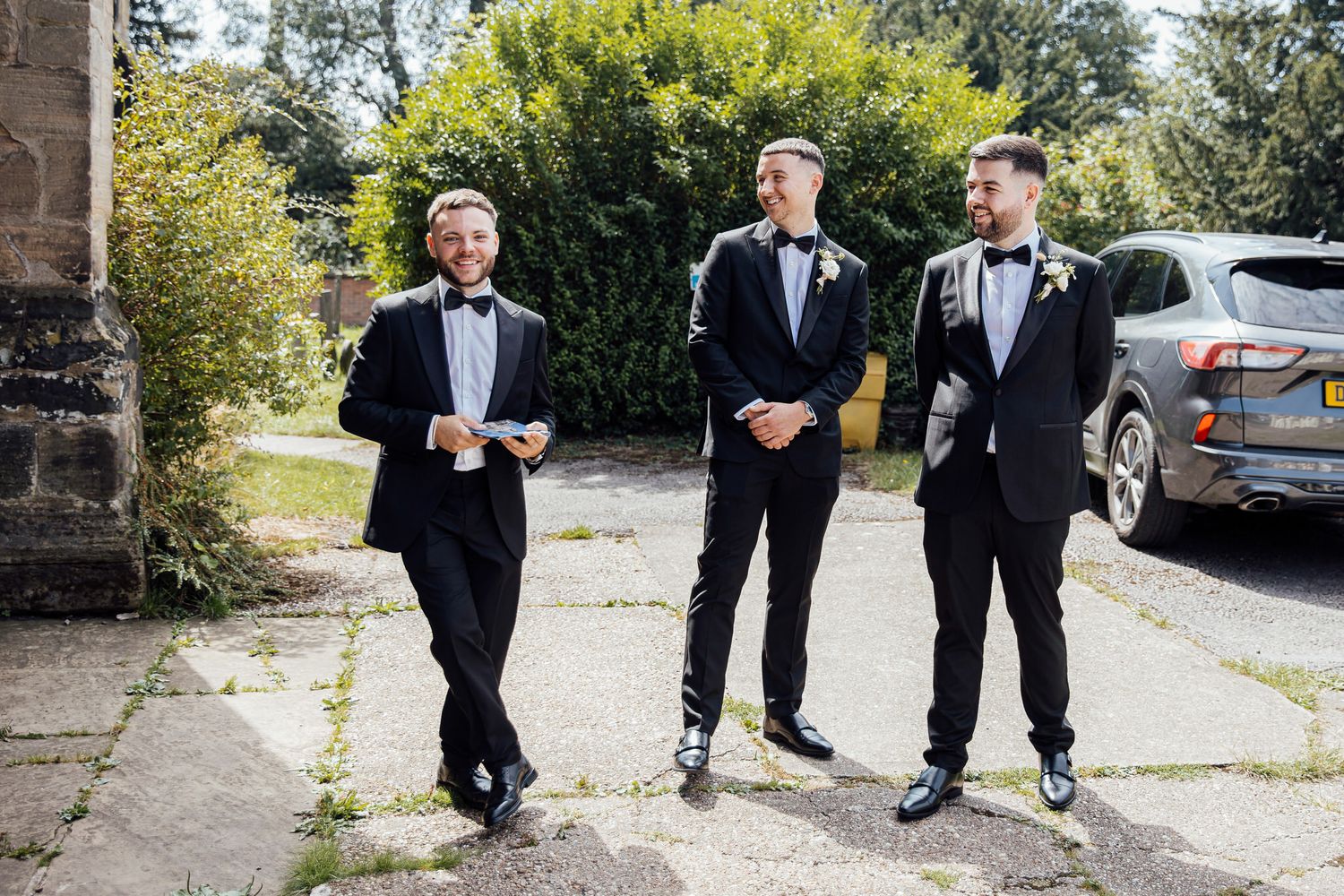 Three groomsmen in black tuxedos standing casually outside stone building.
