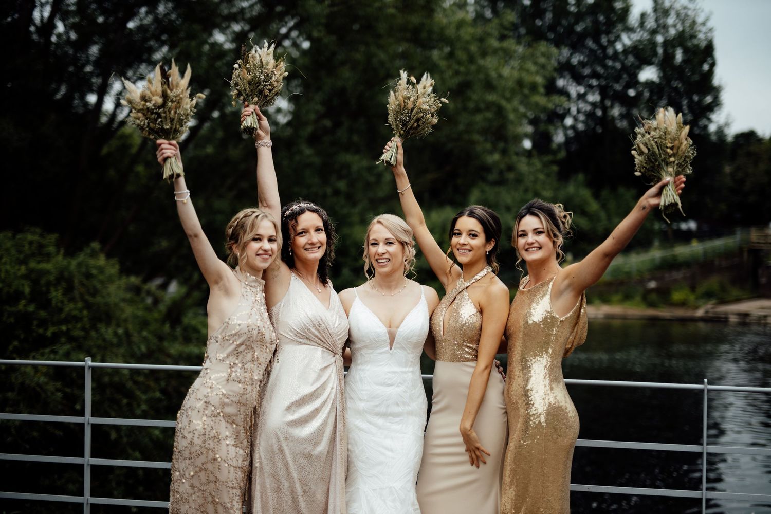 Bridesmaids in champagne dresses celebrate outdoors with bouquets by a waterfront railing.