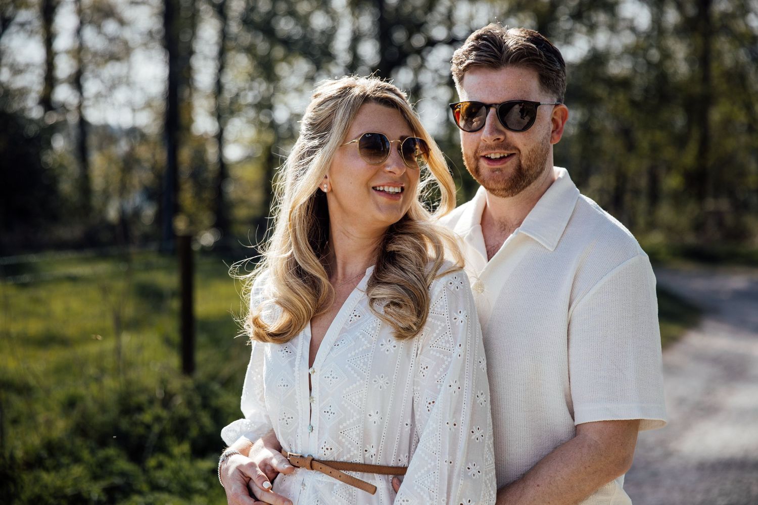 A couple in white and beige outfits share a joyful moment outdoors on a sunny day.