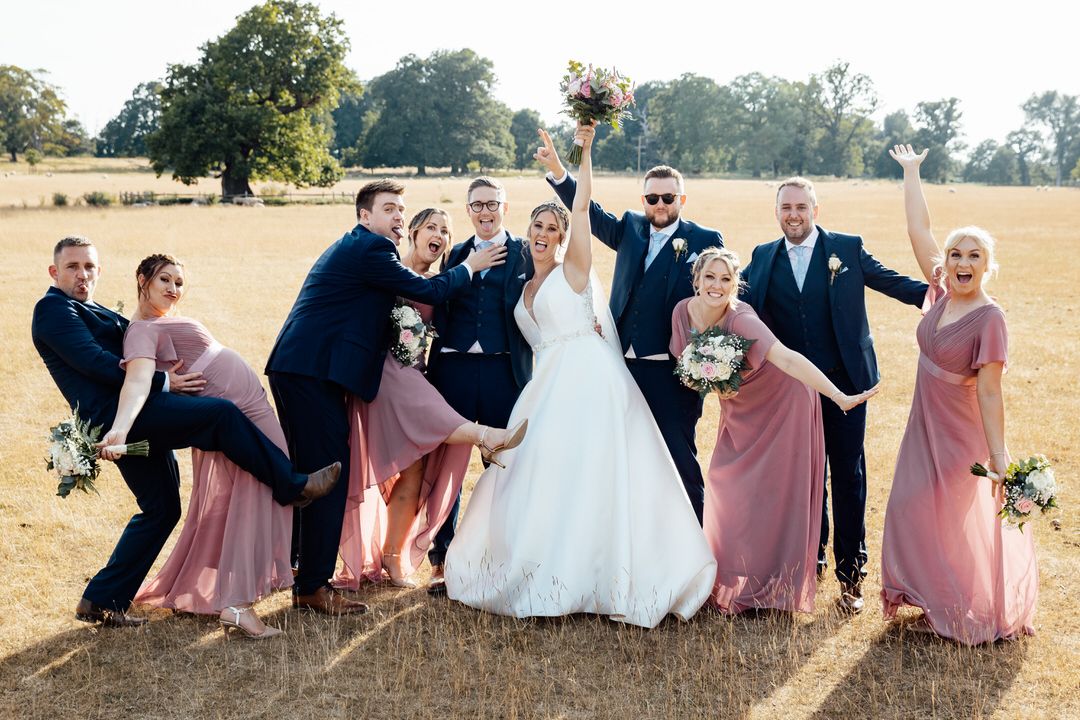 Fun wedding party photo with bridesmaids in pink dresses striking playful poses in outdoor field.