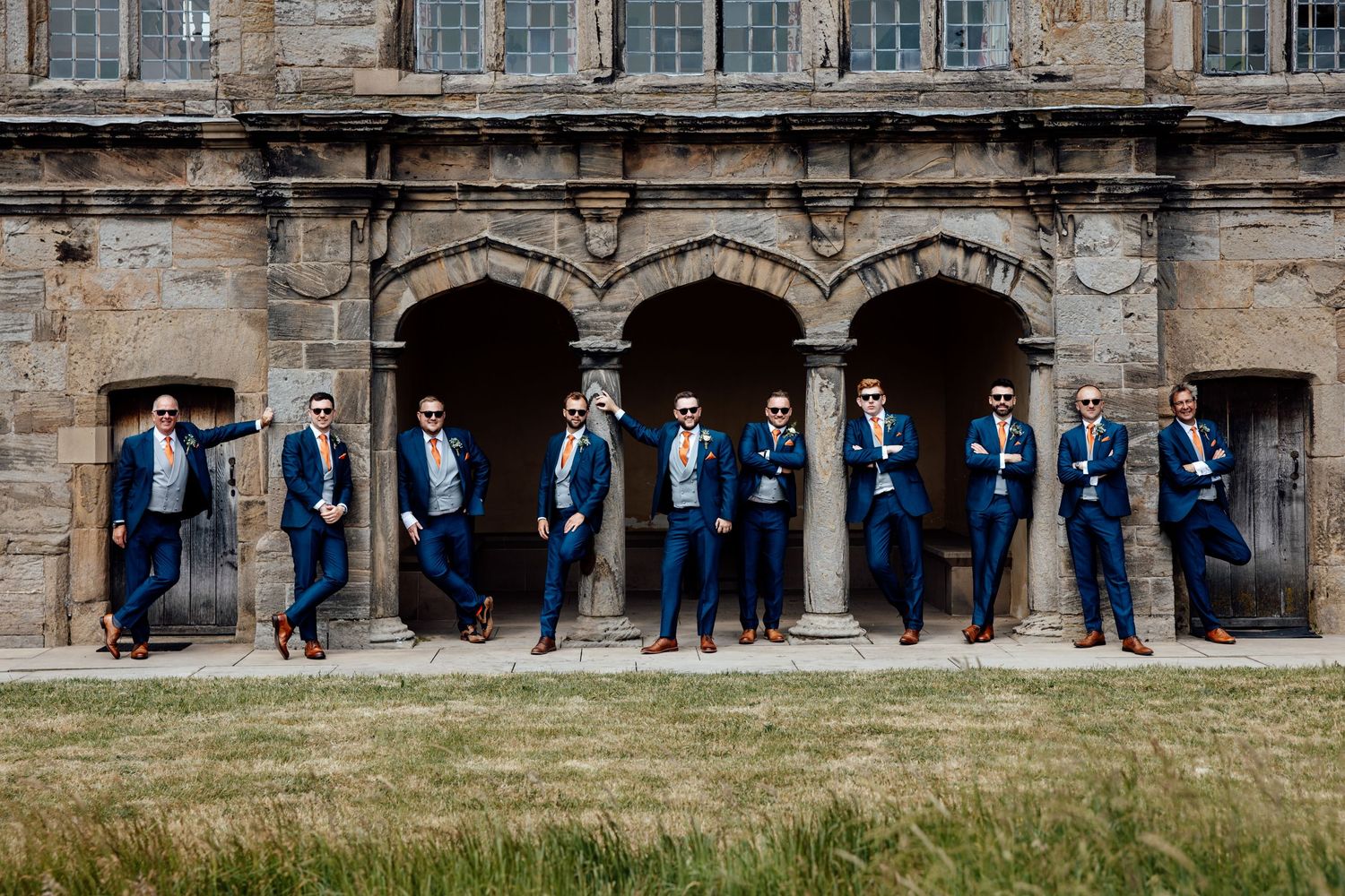 Groomsmen in navy suits posed against stone archway of historic building.