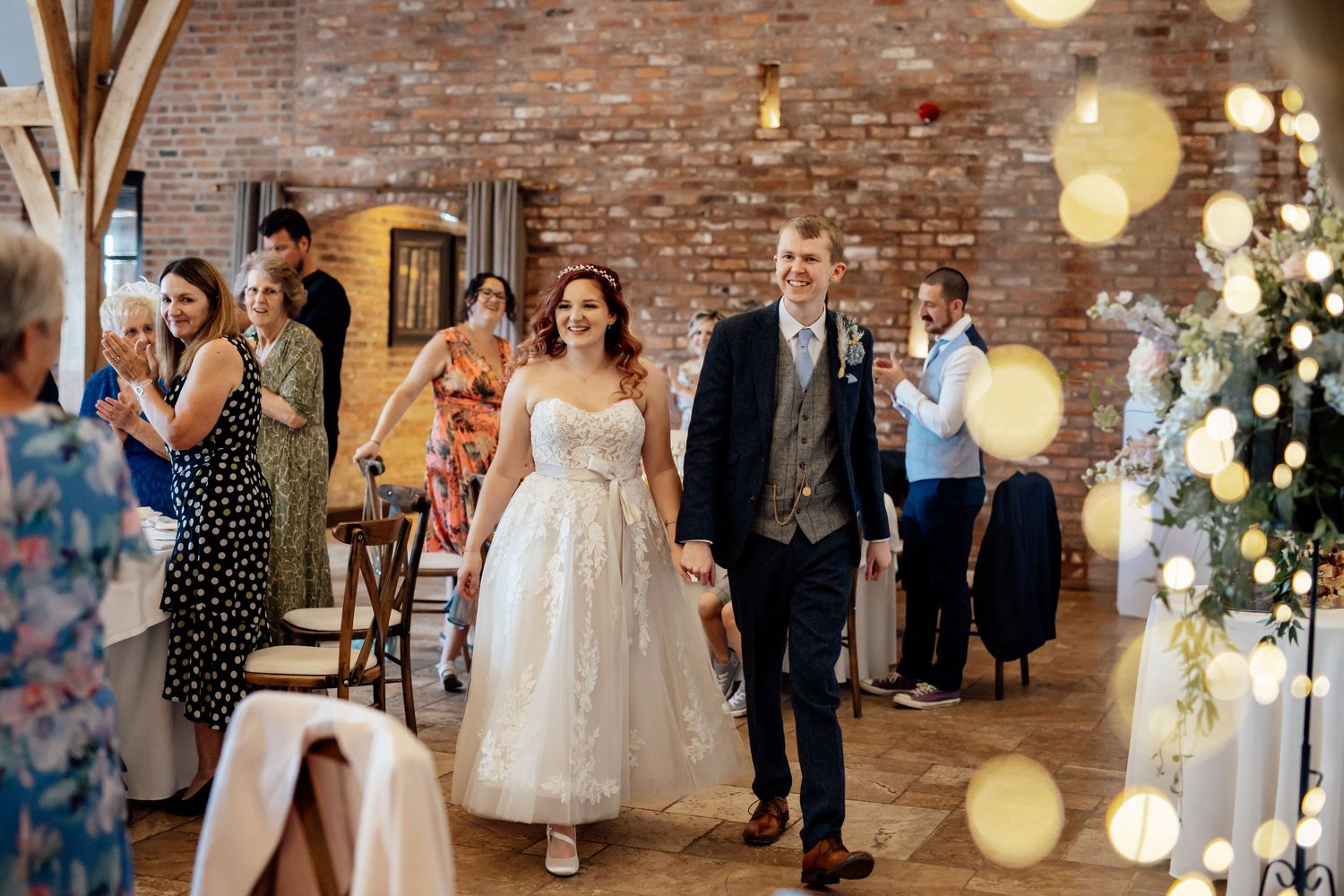 Wedding celebration in a rustic brick venue decorated with hanging lights and floral arrangements.