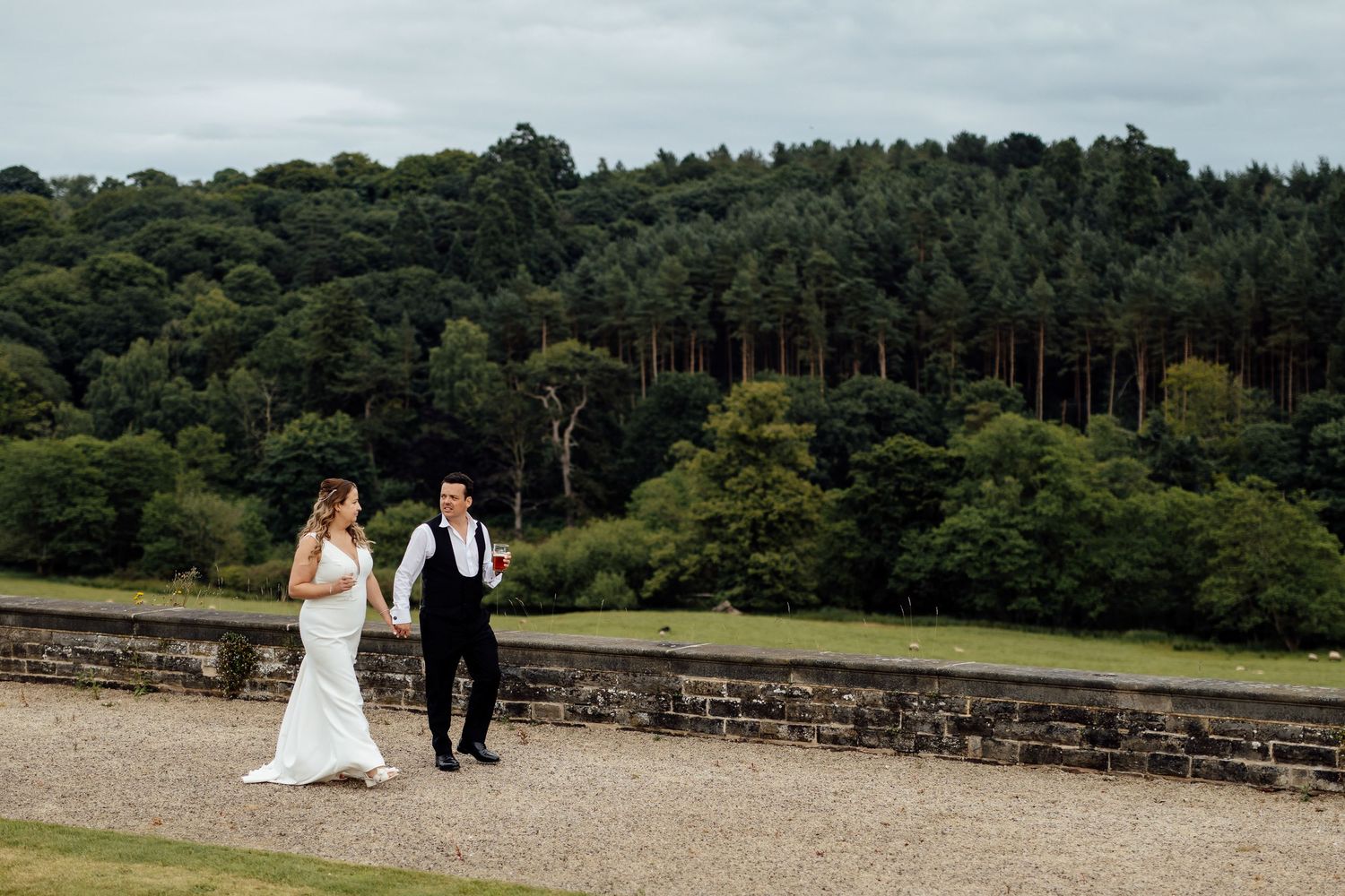 Wedding couple poses together along a scenic stone wall with lush greenery in the background.