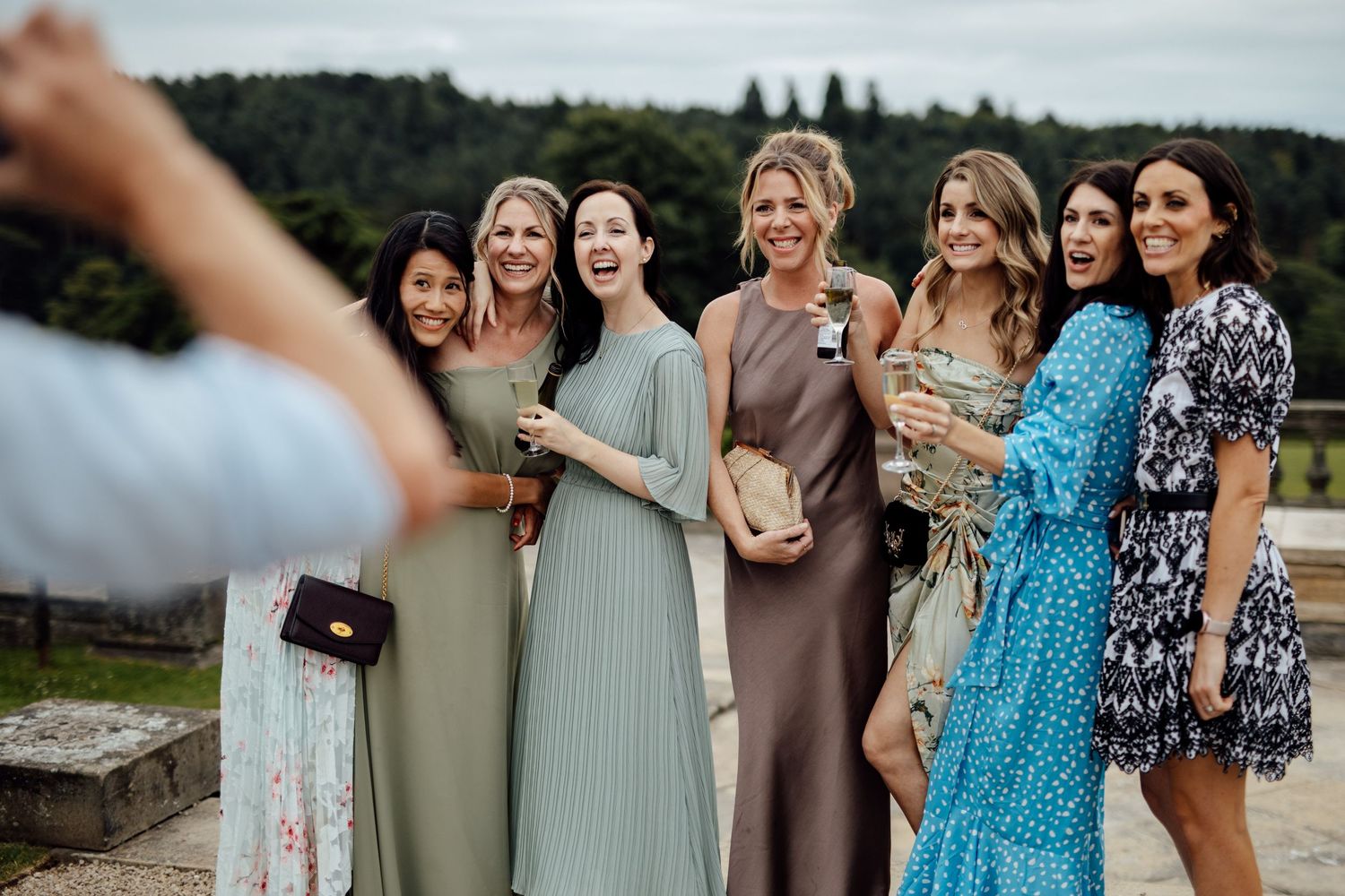 Group of wedding guests in colorful dresses gathered outdoors with scenic view.