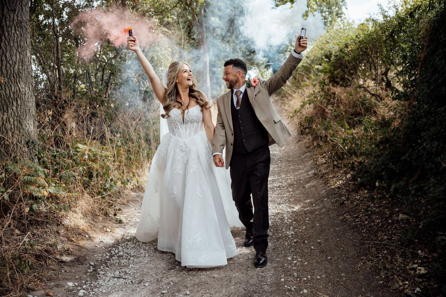 Newlywed couple walking on a forest path holding colorful smoke bombs in wedding attire.