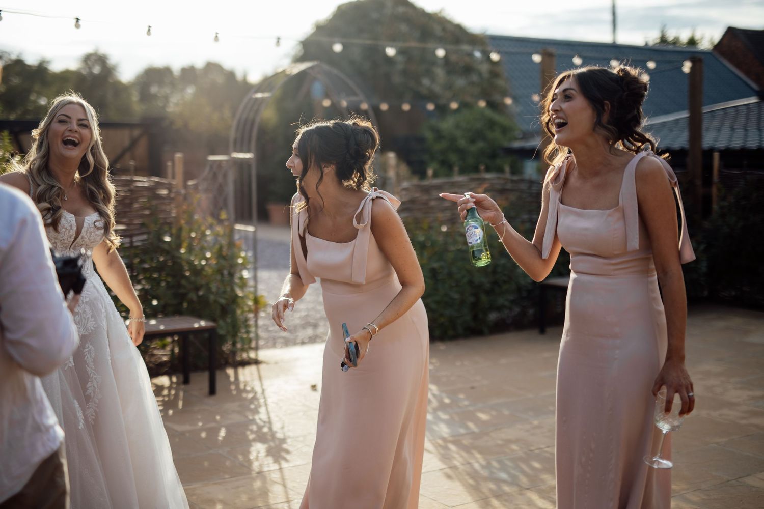 Bridesmaids in blush satin dresses laugh and celebrate outdoors in golden evening wedding reception light.