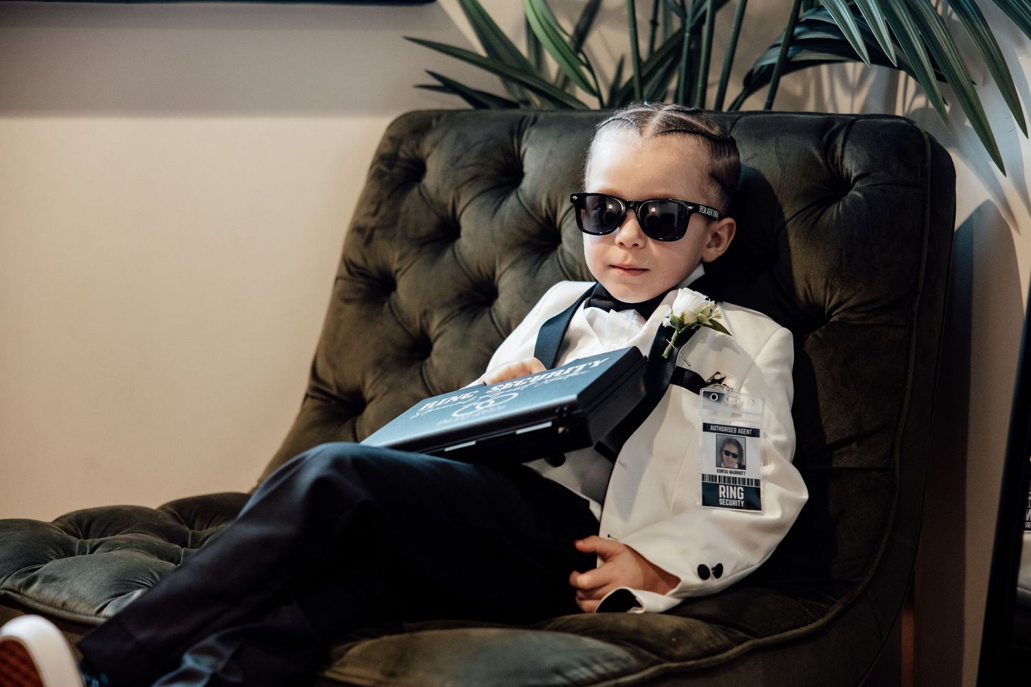 Stylish young ring bearer in sunglasses and tuxedo relaxing in tufted armchair at wedding.