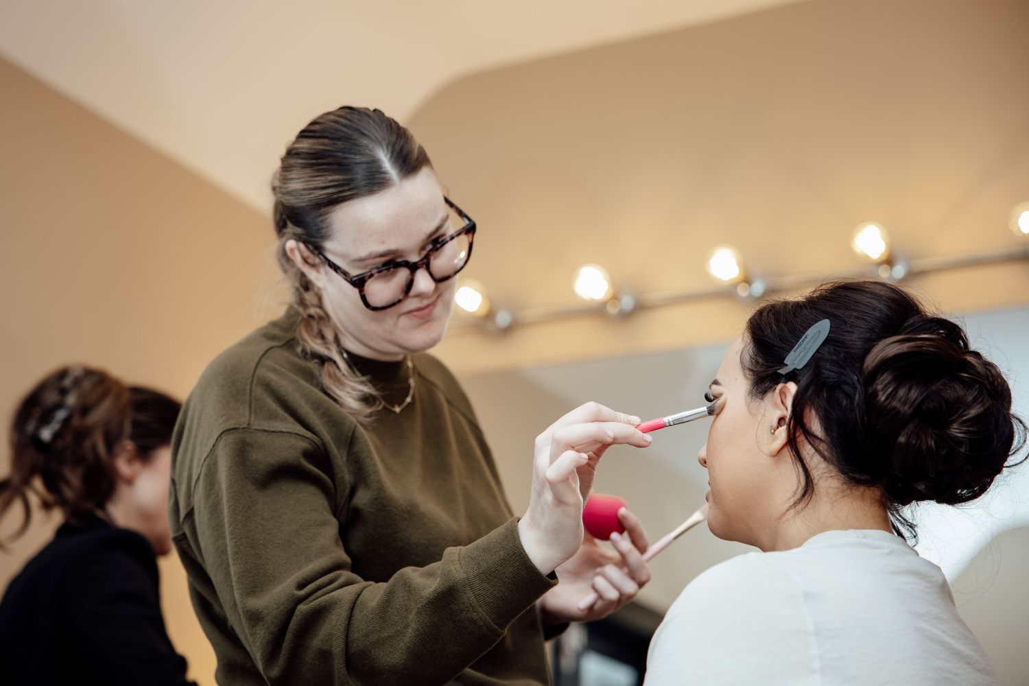 Makeup artist applying cosmetics to a bride with an elegant updo in a professional vanity mirror studio.