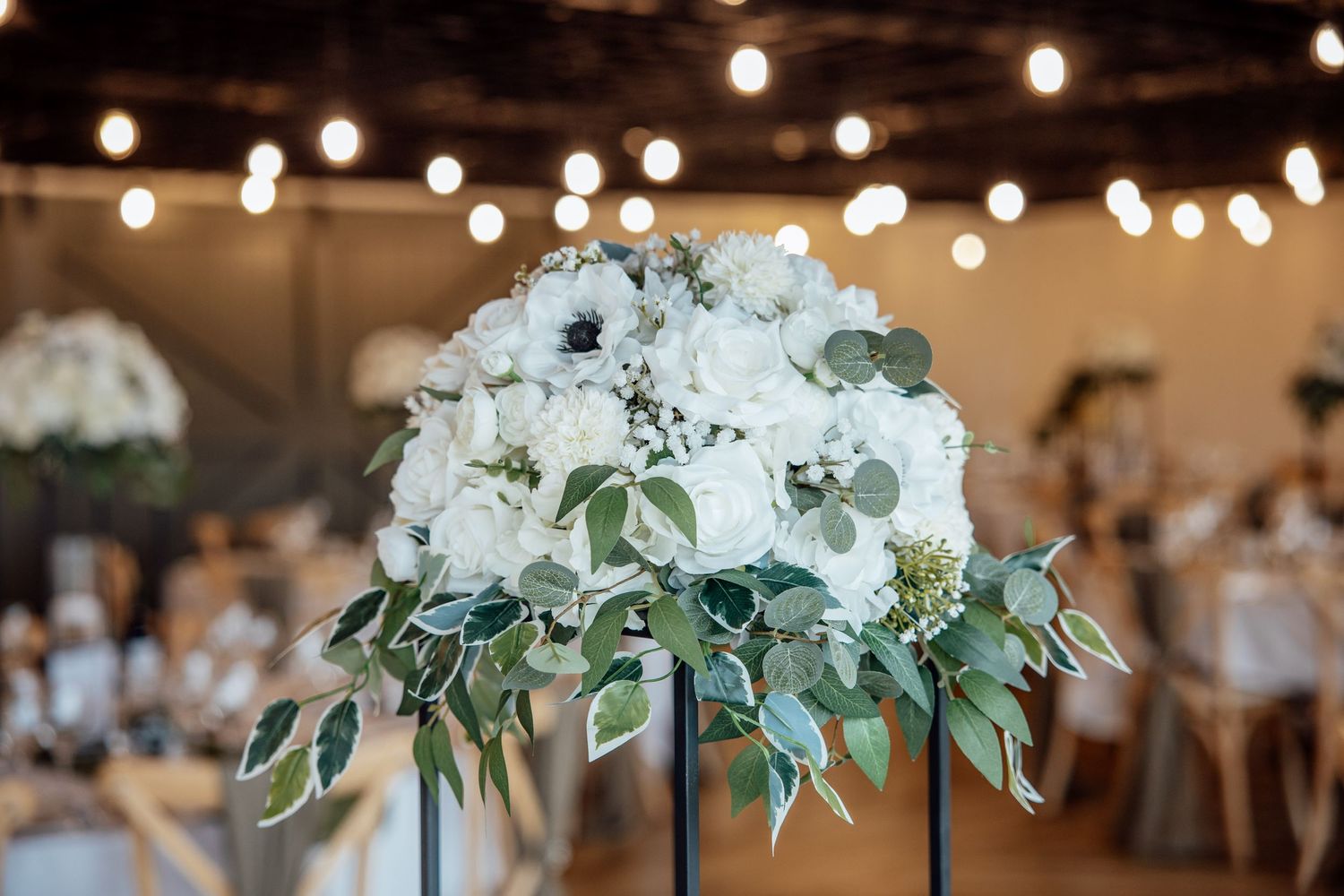 Elegant white floral centerpiece with hydrangeas and eucalyptus under warm string lights at a wedding reception.