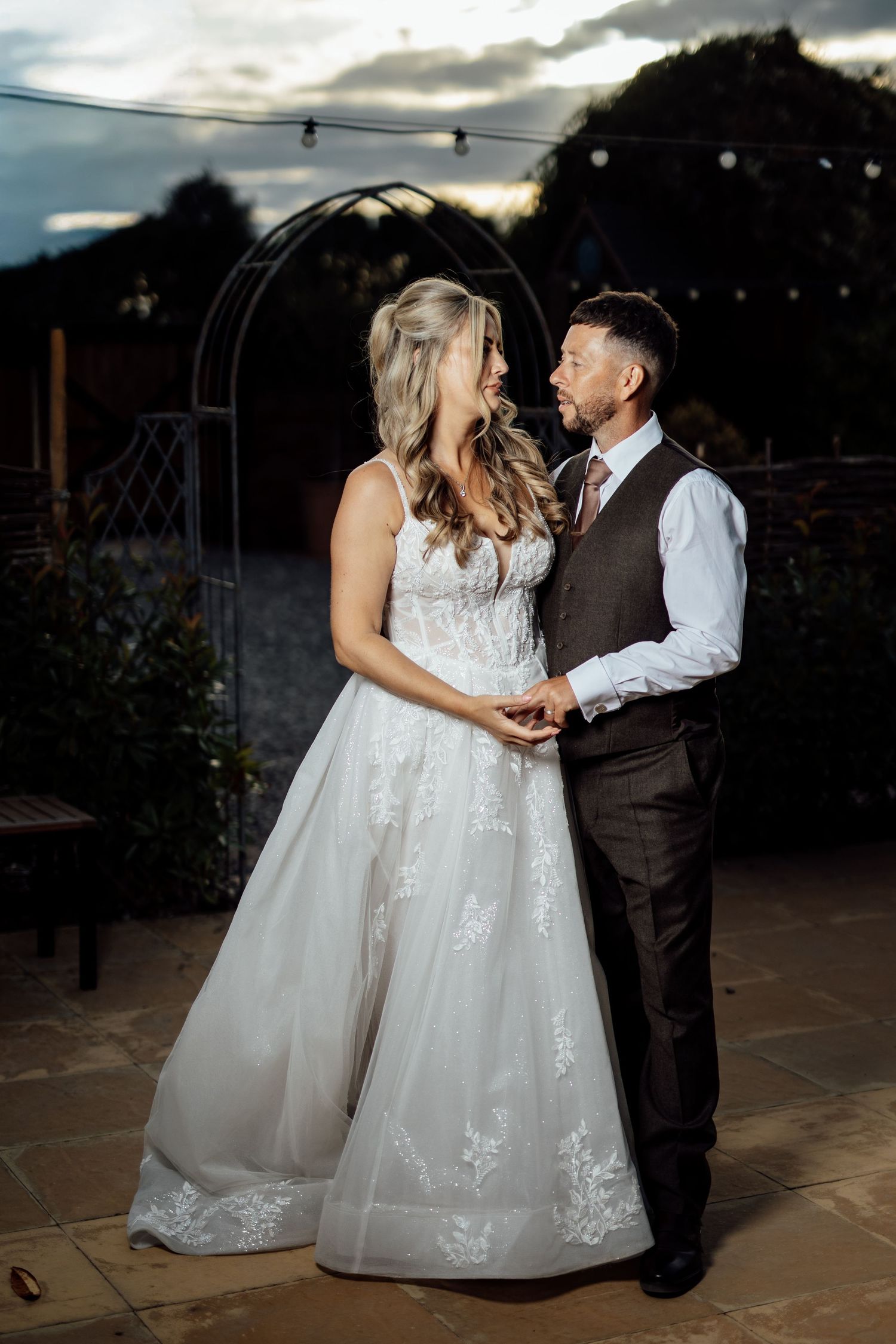 Bride and groom share a romantic moment outdoors at dusk, with string lights and a garden arch backdrop.