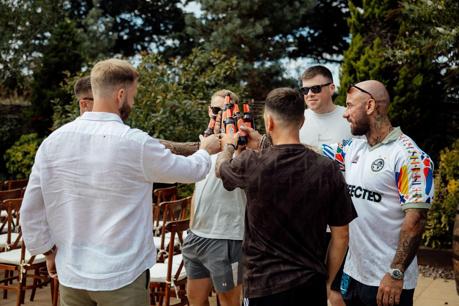 Groomsmen toasting with beer bottles outdoors at a wedding celebration in a garden setting.