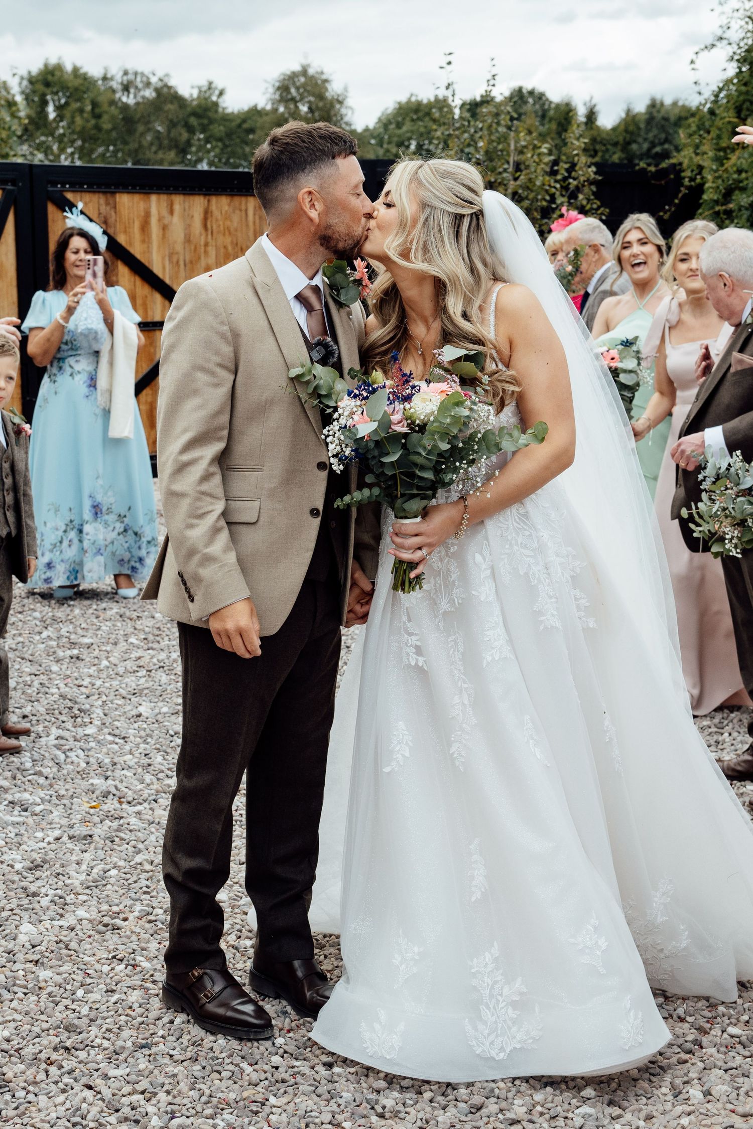 Bride and groom share first kiss outside rustic barn wedding venue with guests celebrating.