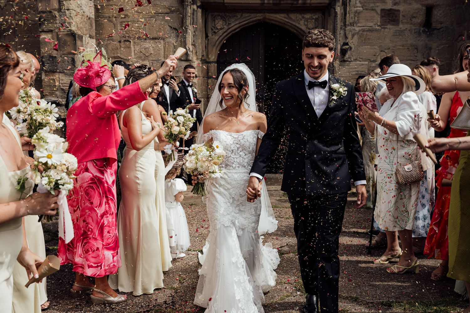 Wedding party celebrates with confetti toss outside historic stone building.