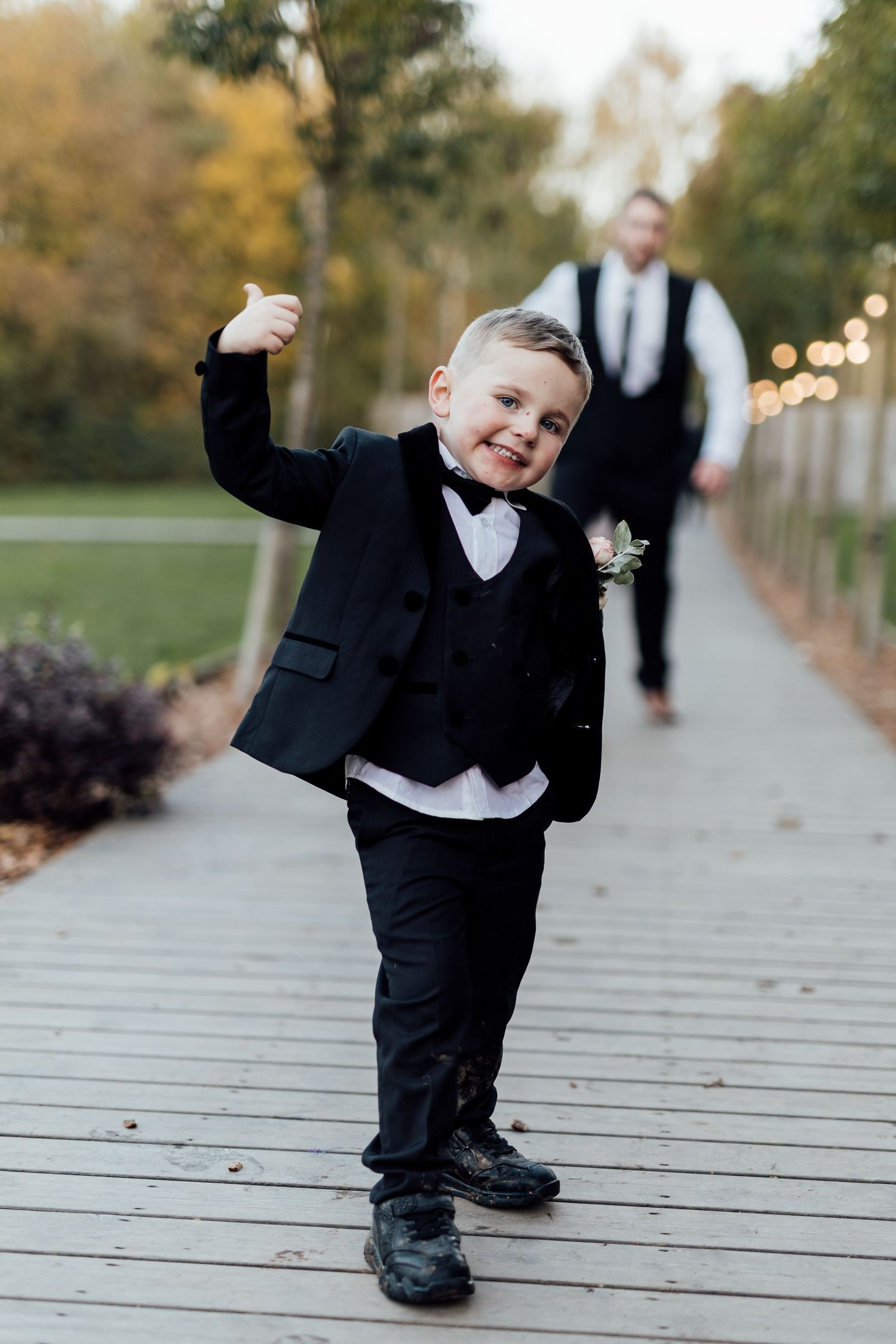 Young ring bearer in formal black tuxedo strikes playful pose on wooden walkway during outdoor wedding.