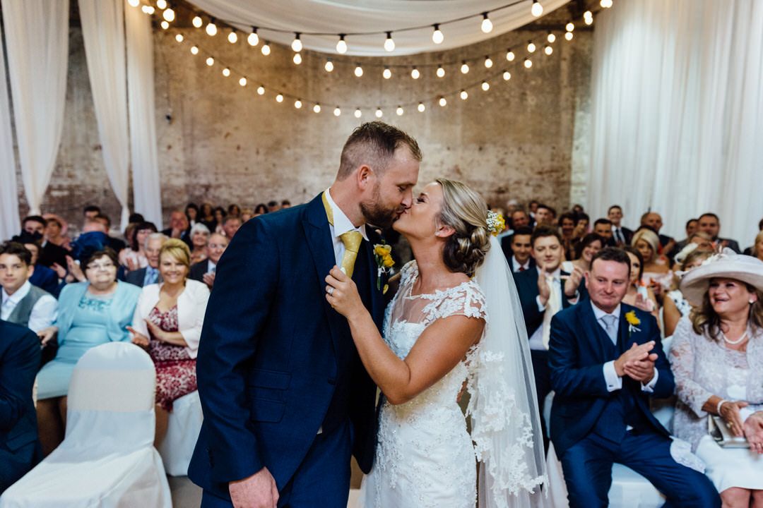A romantic wedding ceremony under string lights with guests watching in an industrial venue.