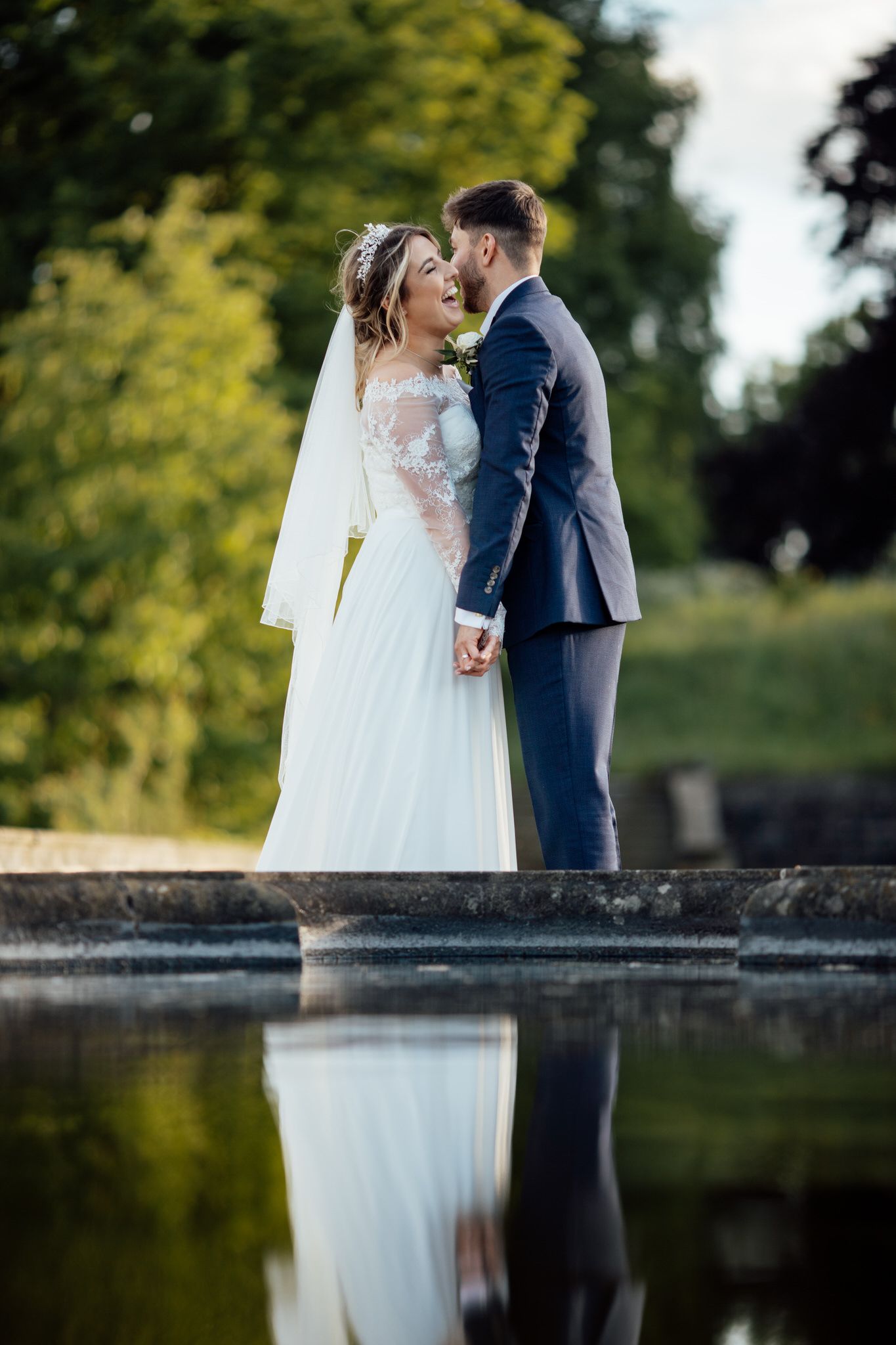 Elegant wedding couple poses by reflective water with lush greenery in background.