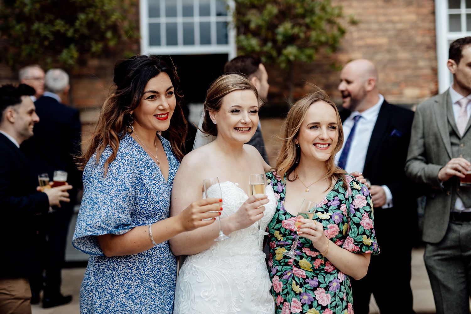 Wedding guests mingling outdoors near a brick building on a sunny day.