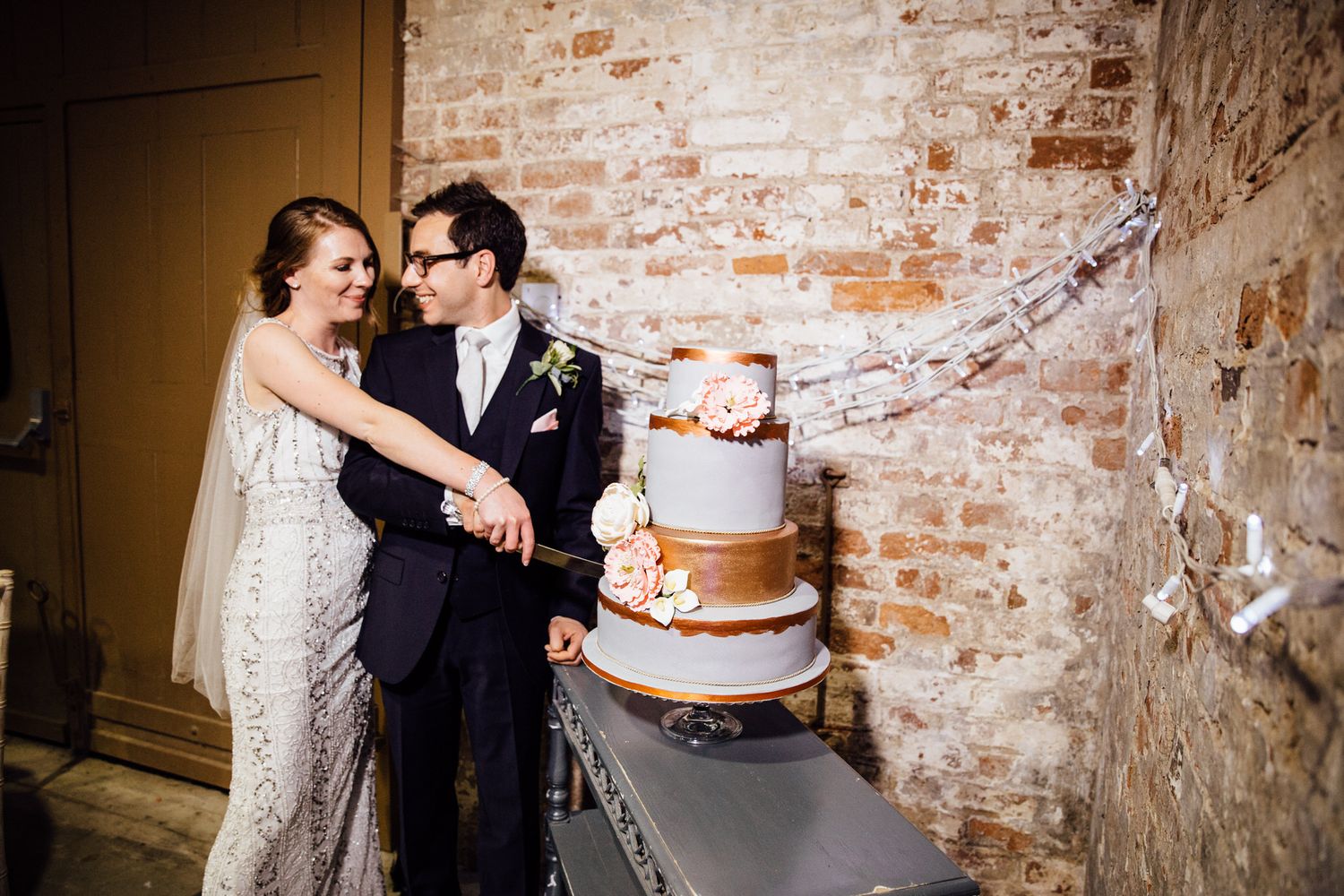 Couple cutting their wedding cake together in front of an exposed brick wall.