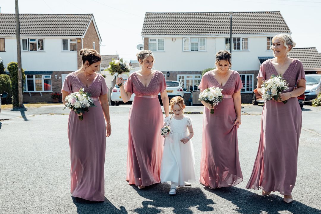 Bridesmaids in dusty rose dresses gather outside on a sunny day holding white floral bouquets.