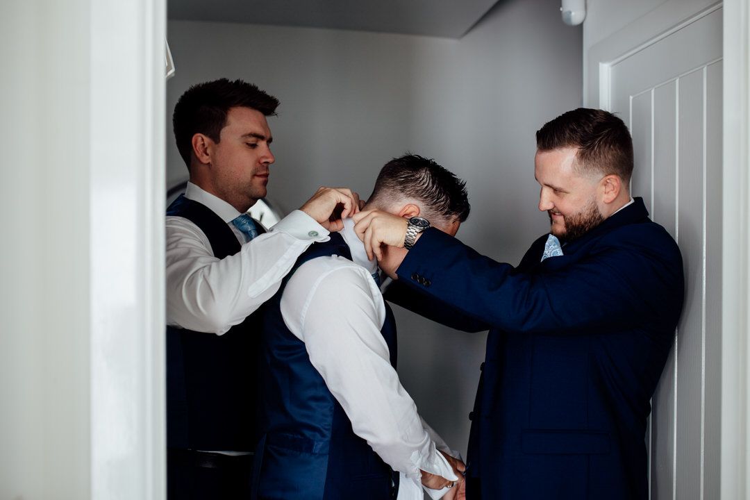 Groomsmen help each other get dressed in navy suits before wedding ceremony.