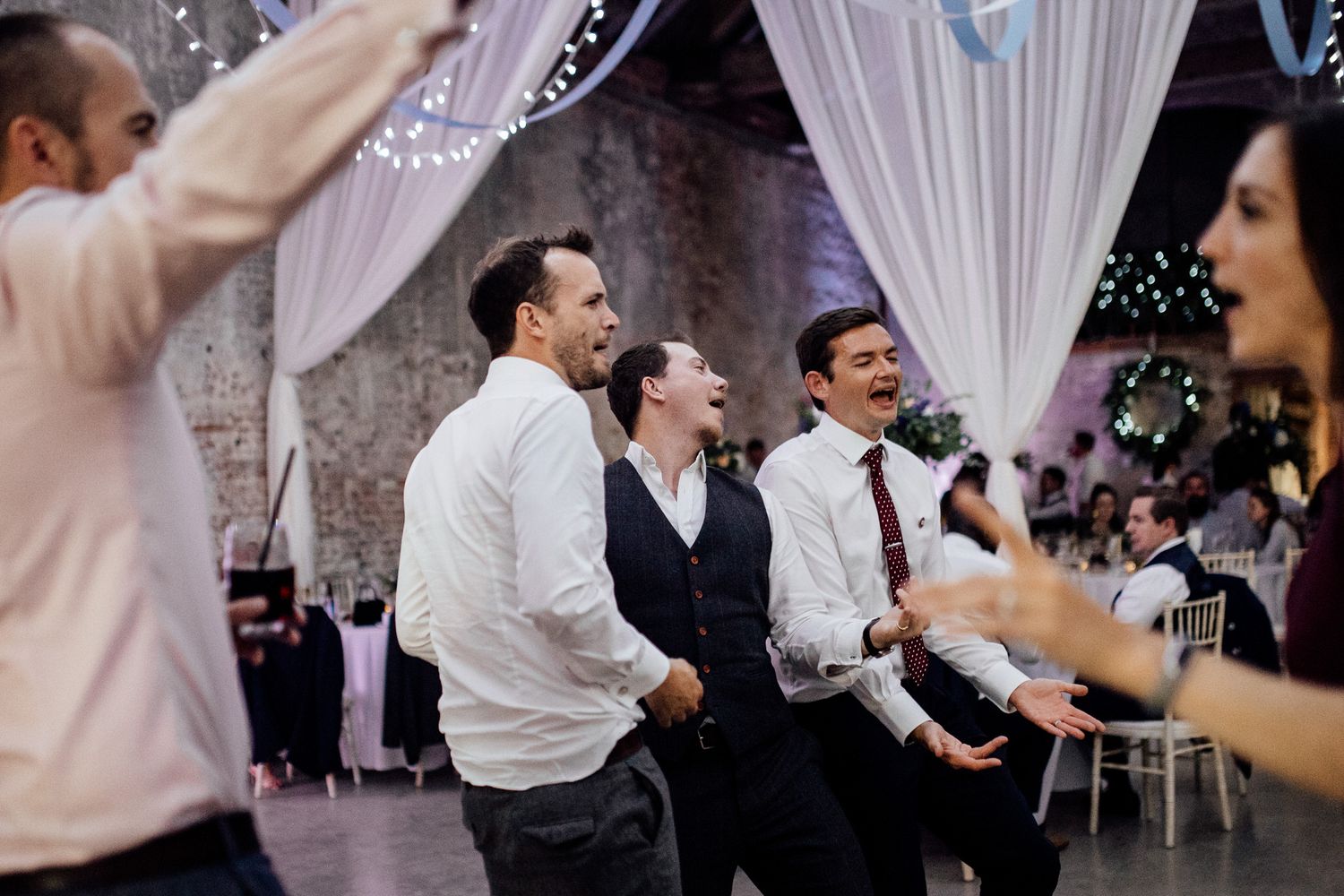 Wedding guests dancing and celebrating on a dance floor with white drapes in the background.