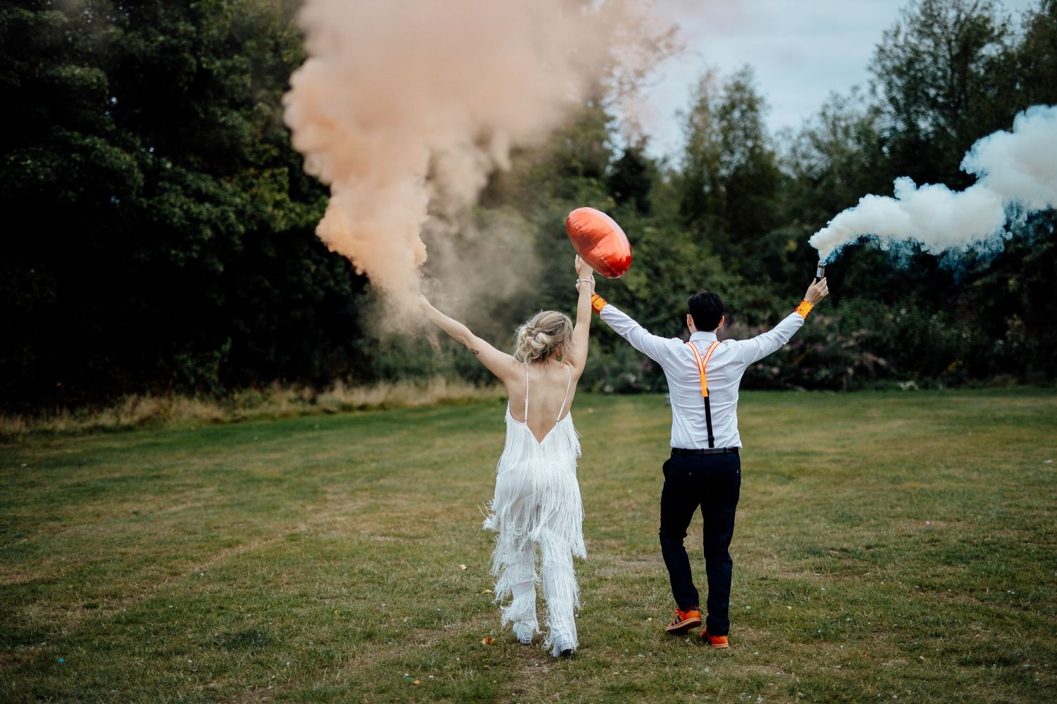 Newlyweds celebrate with colorful smoke bombs in an outdoor field.