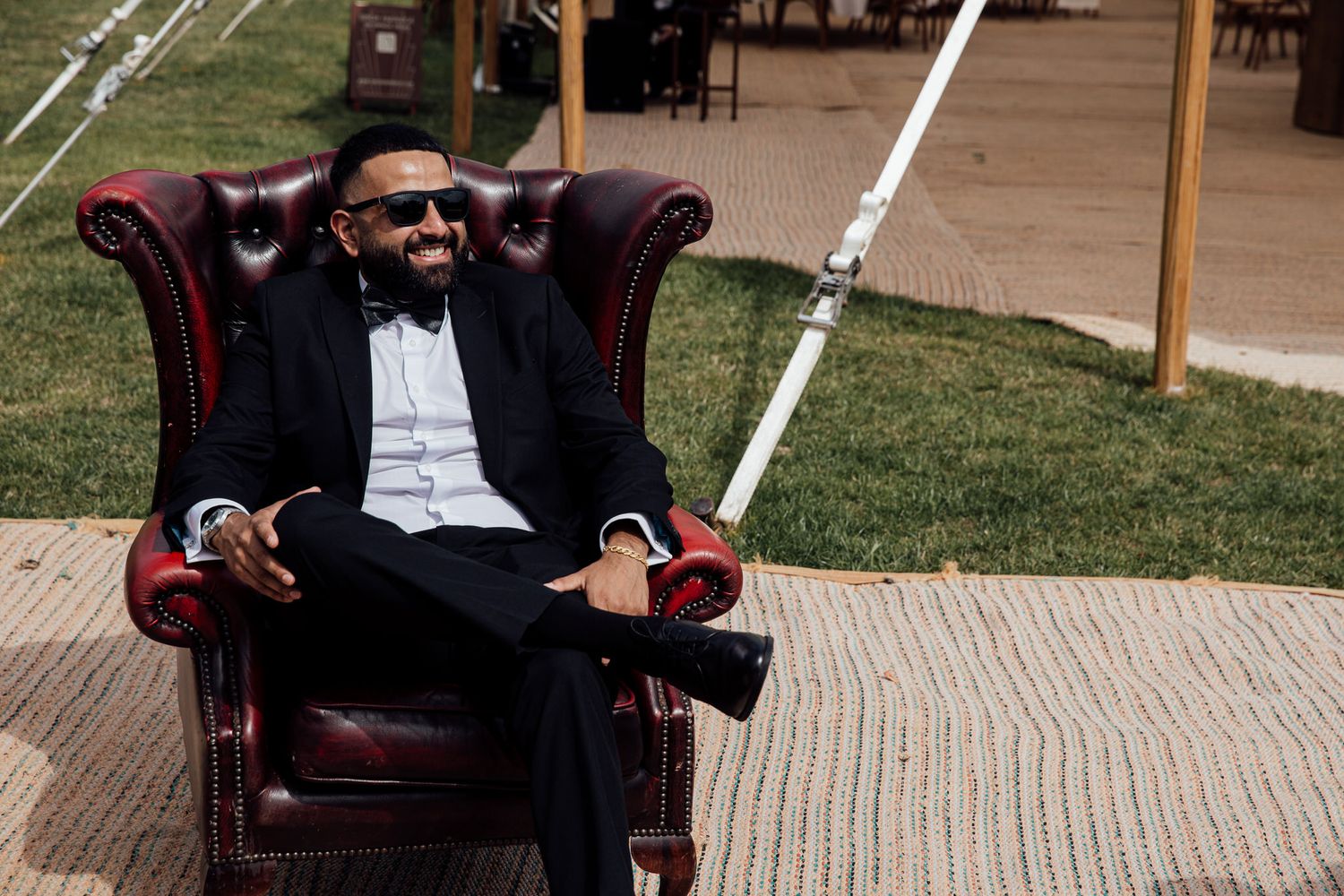 A stylishly dressed guest relaxes in a leather armchair at an outdoor wedding venue.