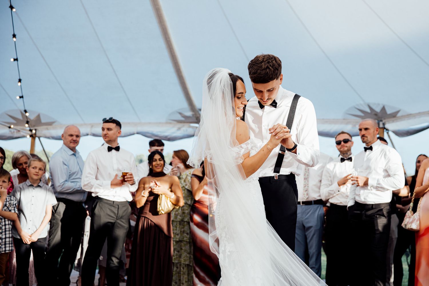 A couple shares their first dance at an outdoor wedding reception under a tent as guests look on.