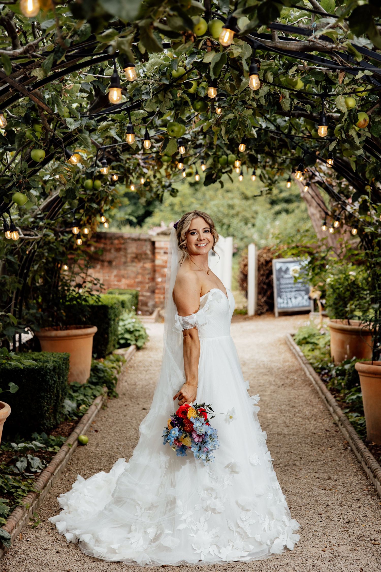 String lights illuminate a garden path with terracotta pots and greenery.