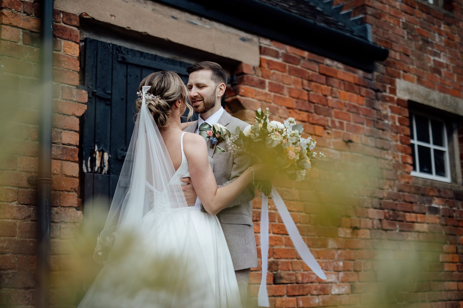 A romantic wedding couple poses against a rustic brick wall backdrop.