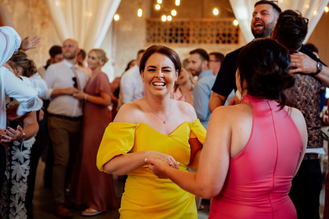 Wedding guests dance and celebrate on a rustic venue dance floor under warm lighting and white drapes.
