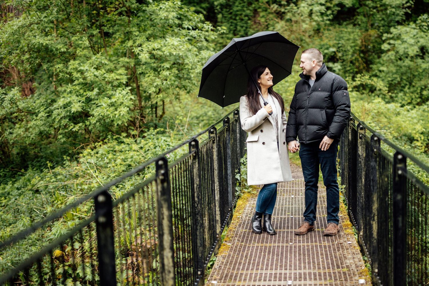 Two people stroll hand in hand across a metal bridge surrounded by dense forest foliage.