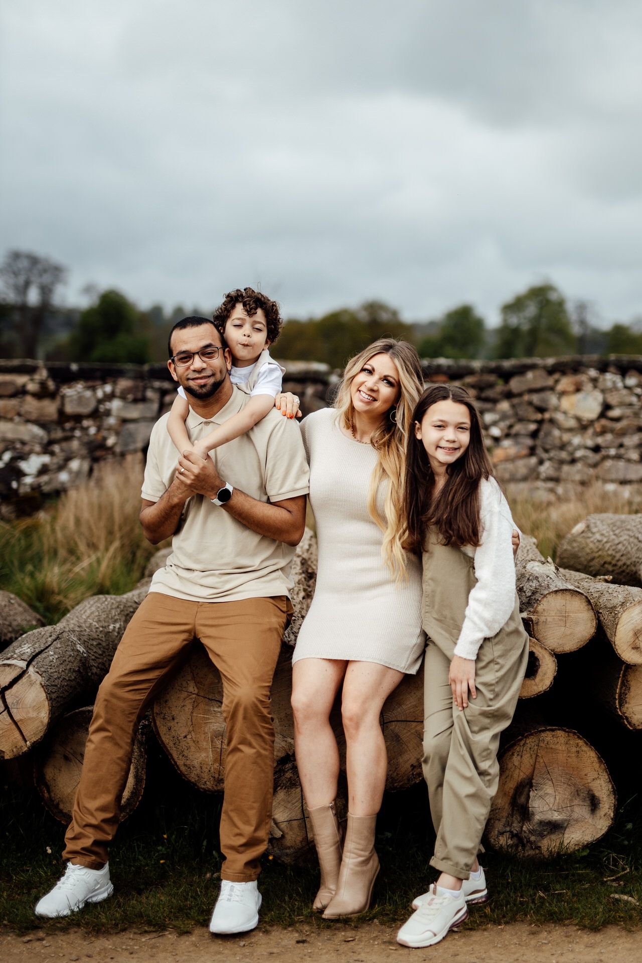 A family in coordinated neutral outfits poses casually against rustic wooden logs in an outdoor setting.
