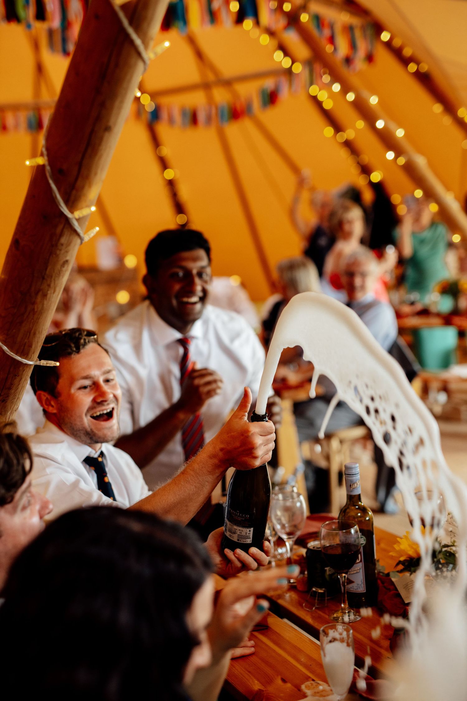 Guests celebrating with champagne spray at a festive wedding reception under an orange tent.