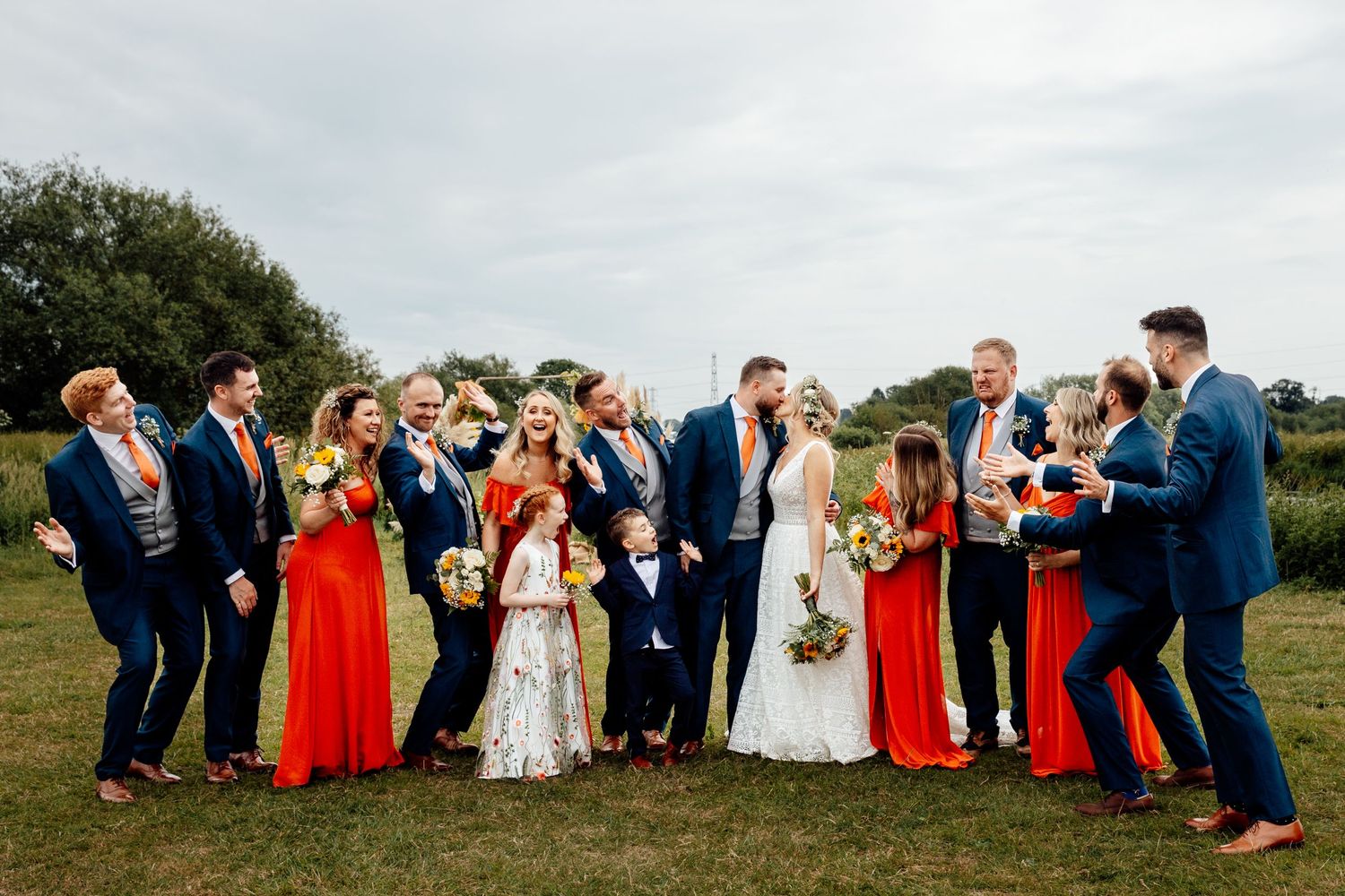 Wedding party group photo with guests in blue suits and red dresses on a grassy hill.