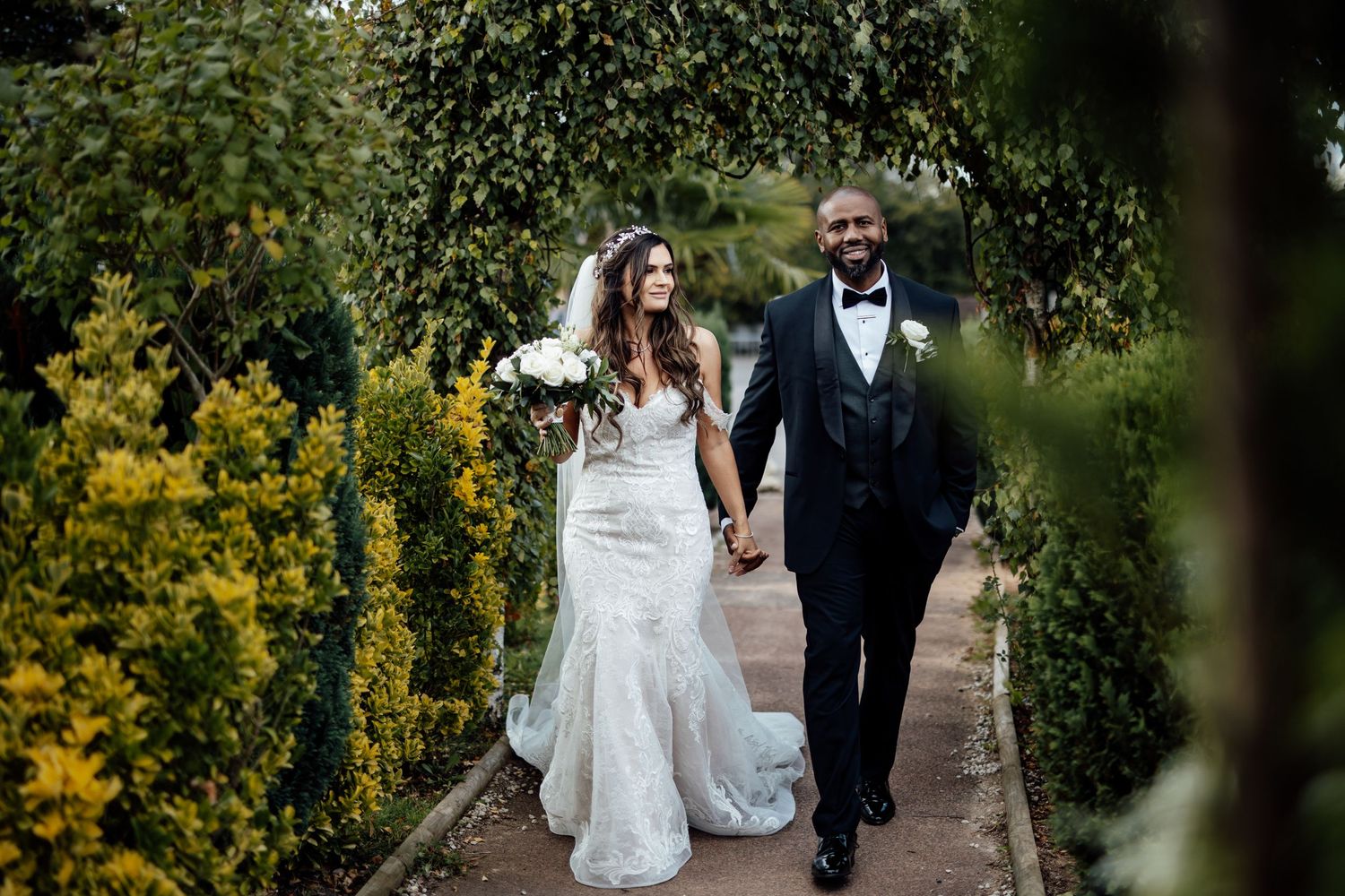 Newlyweds walking hand in hand through a garden path lined with yellow flowers and greenery.