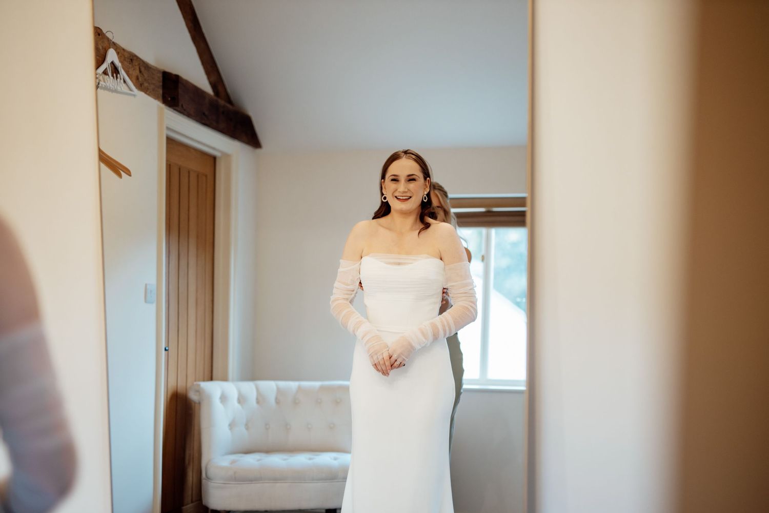 A bride in a white wedding gown stands in a sunlit room during final preparations.