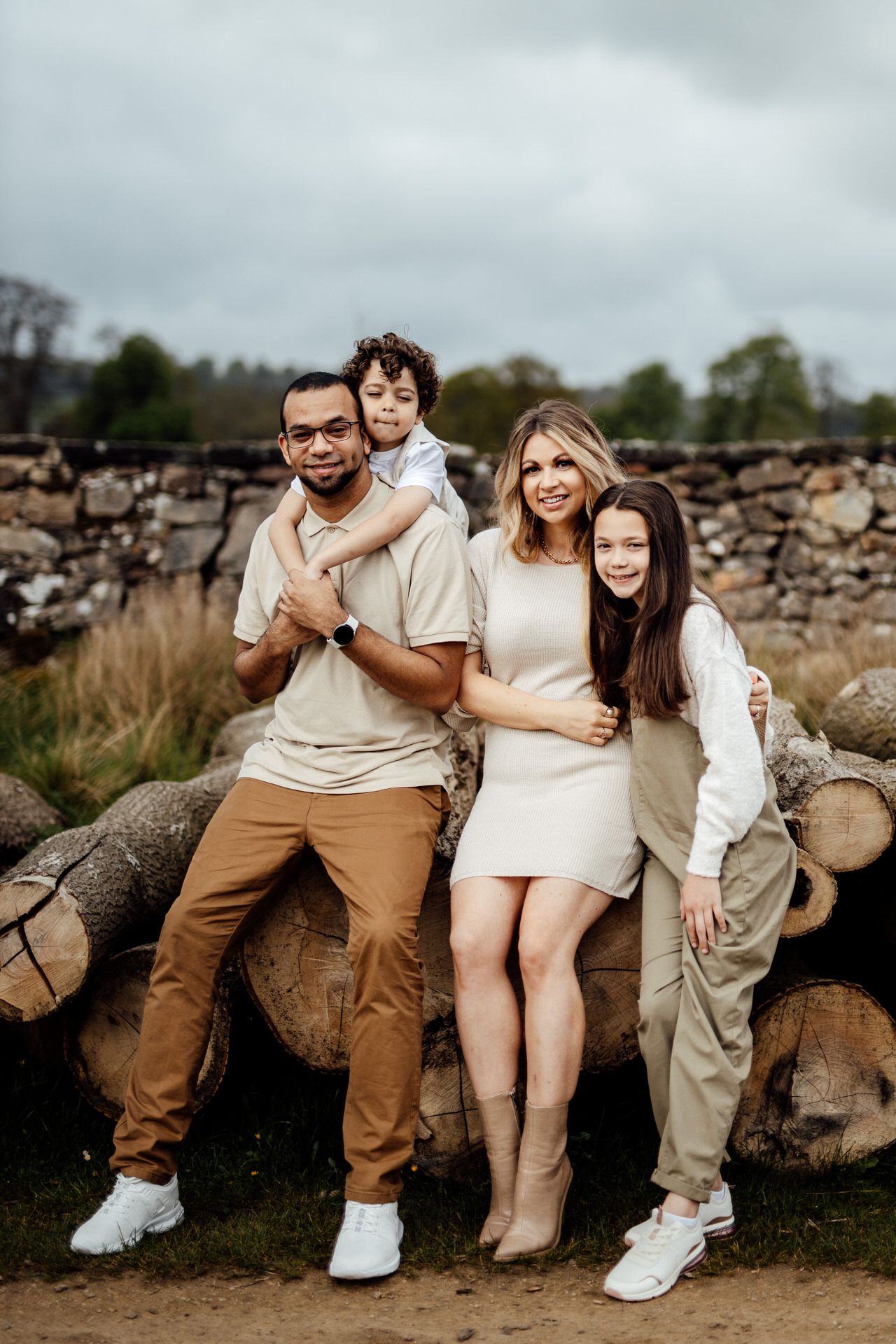 Casual family portrait session on rustic wooden logs against a cloudy sky backdrop.