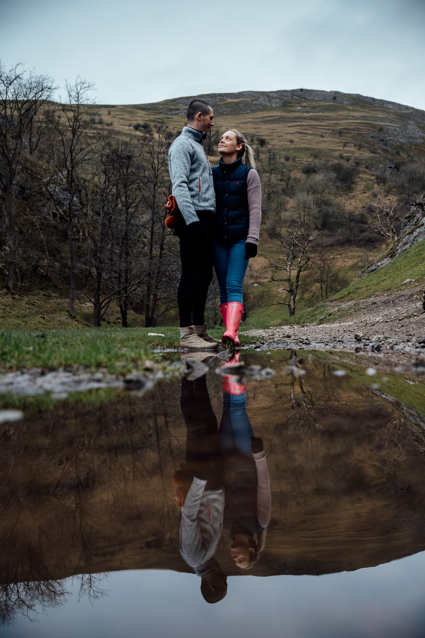 A romantic couple stands by a reflective pond in the mountains, their image mirrored in the still water.