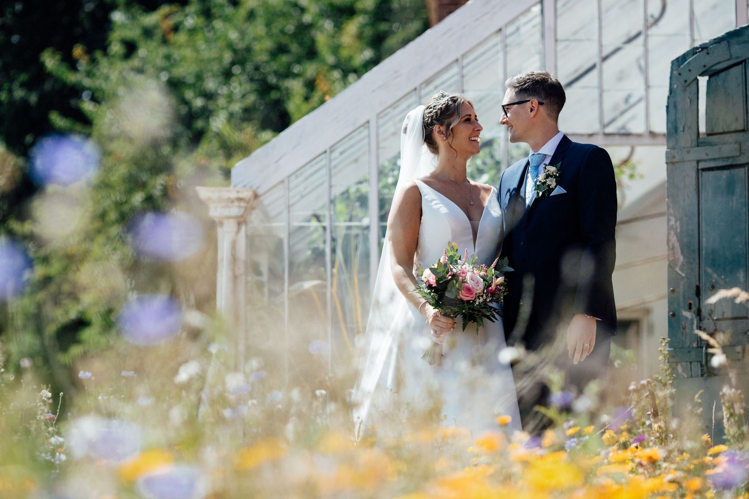 A couple in wedding attire stands on outdoor stairs surrounded by colorful summer flowers and greenery.