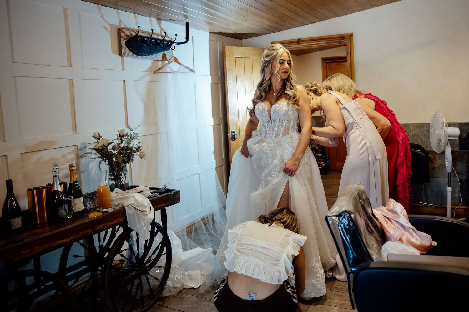 Bride in white lace gown being helped by bridesmaids while getting ready in a rustic bridal suite.