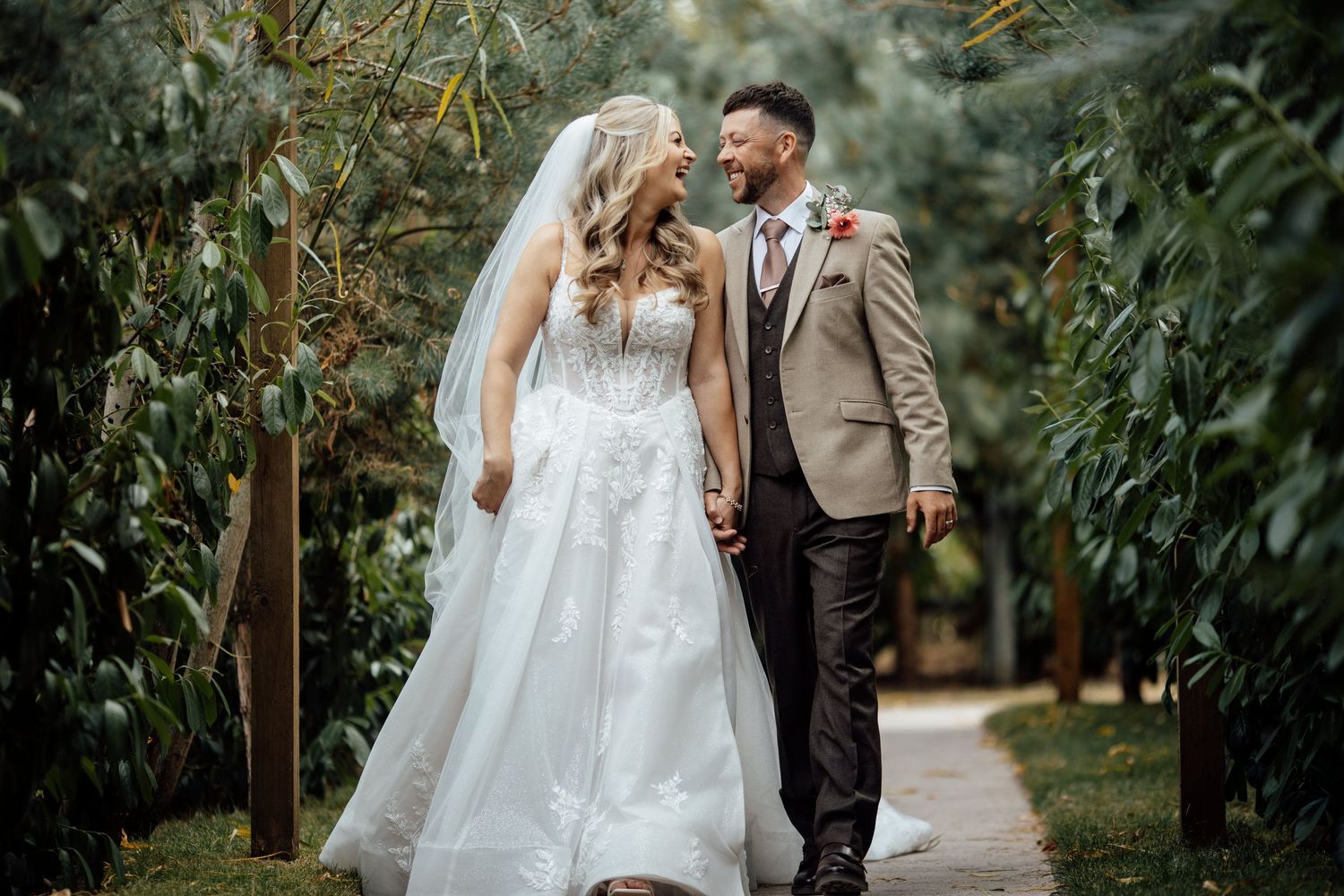 Bride and groom walk hand in hand through lush green garden pathway on their wedding day.