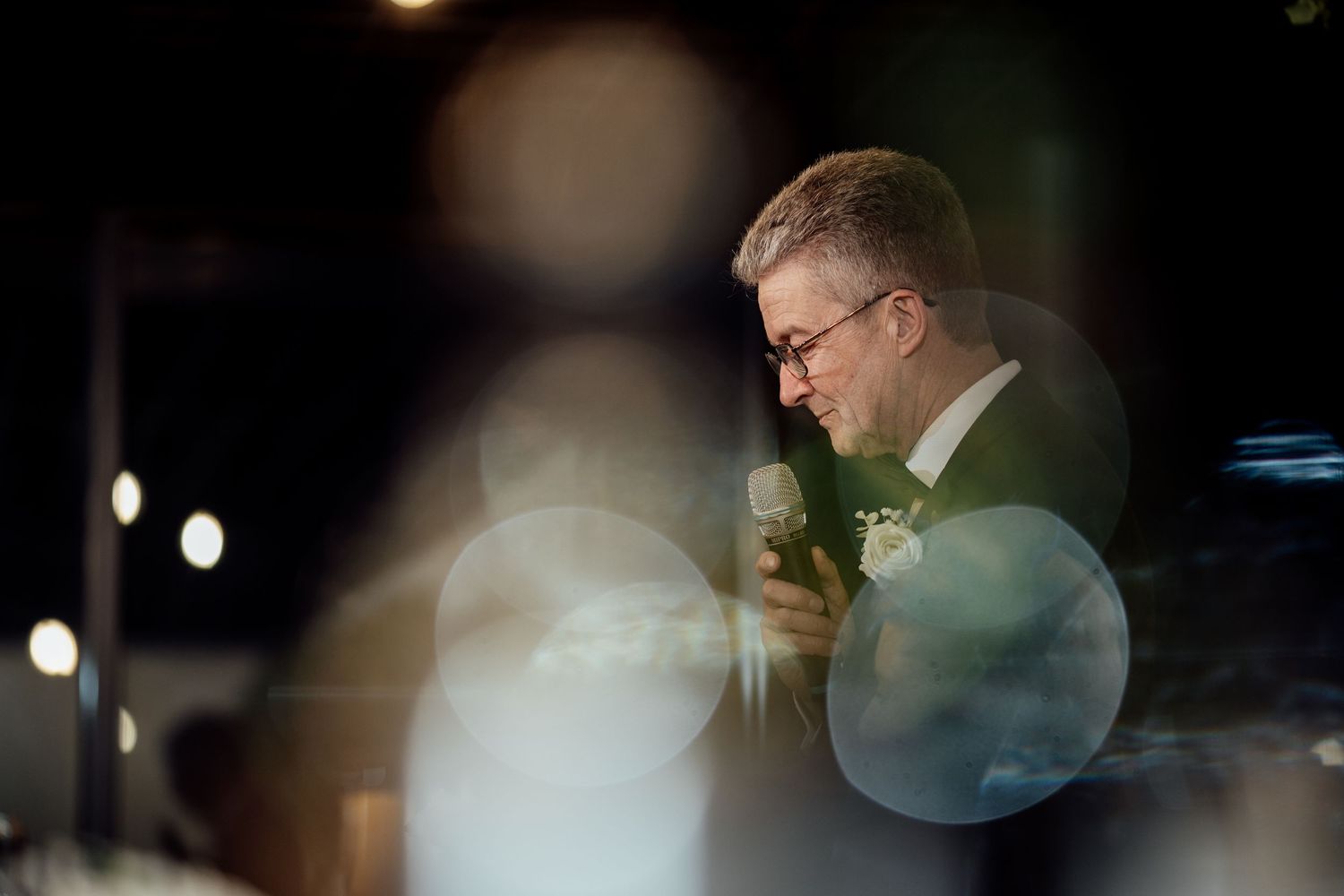 Emotional man in suit delivers heartfelt wedding speech under soft bokeh lights at evening reception.