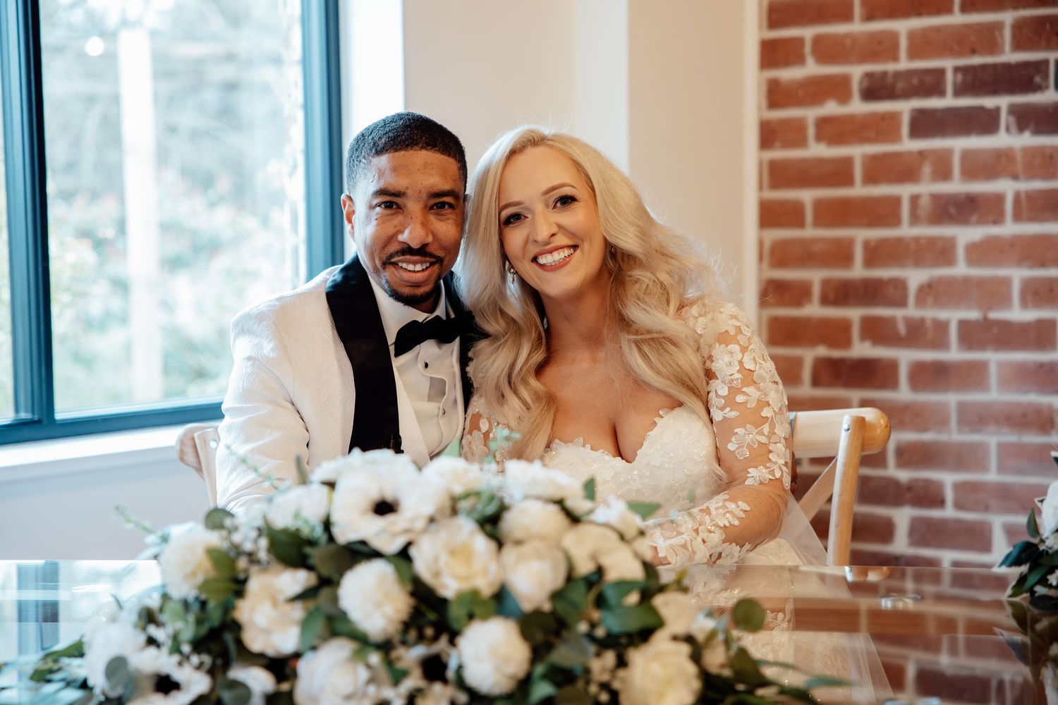 Smiling bride and groom in white tuxedo seated at wedding reception table with white floral centerpiece.