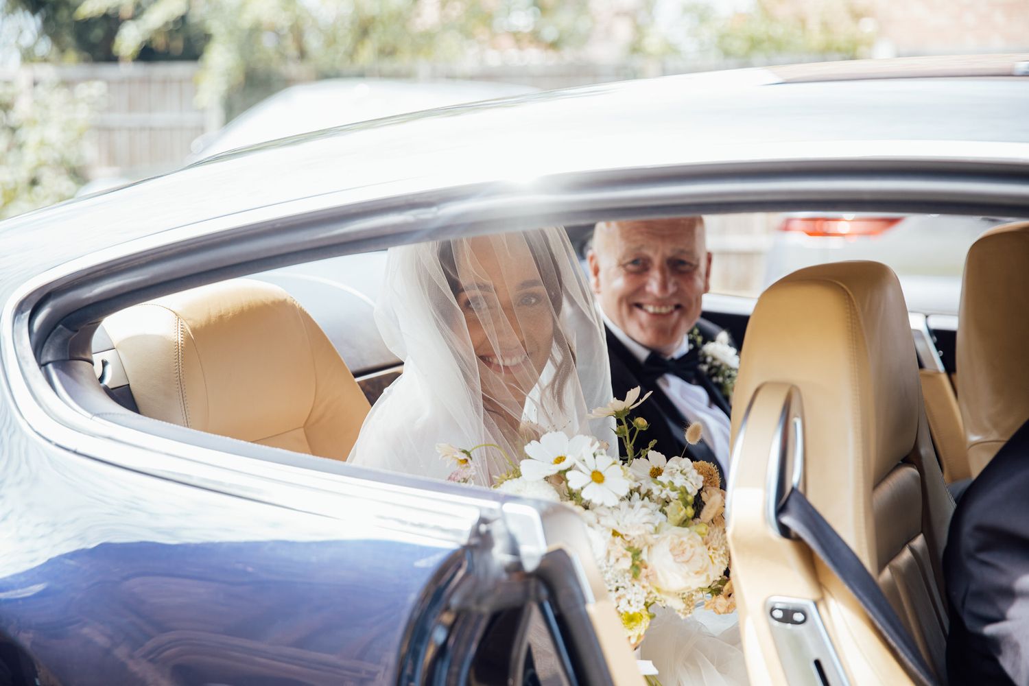 Wedding couple sits in luxury car with elegant interior and floral bouquet.