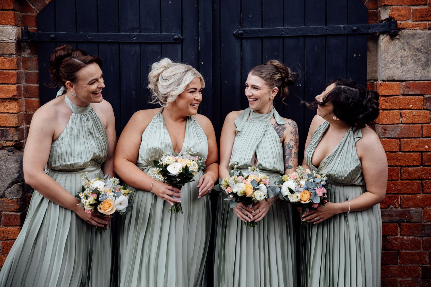 Bridesmaids in sage green dresses stand together holding bouquets against a brick wall backdrop.