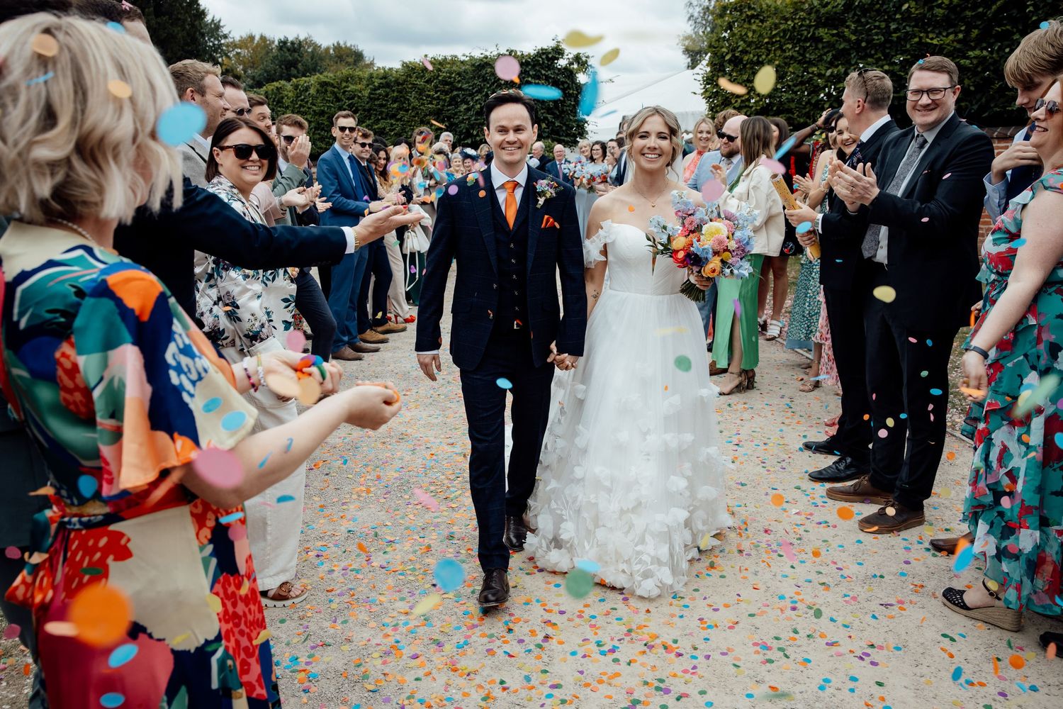 Wedding guests throw colorful confetti during an outdoor celebration.