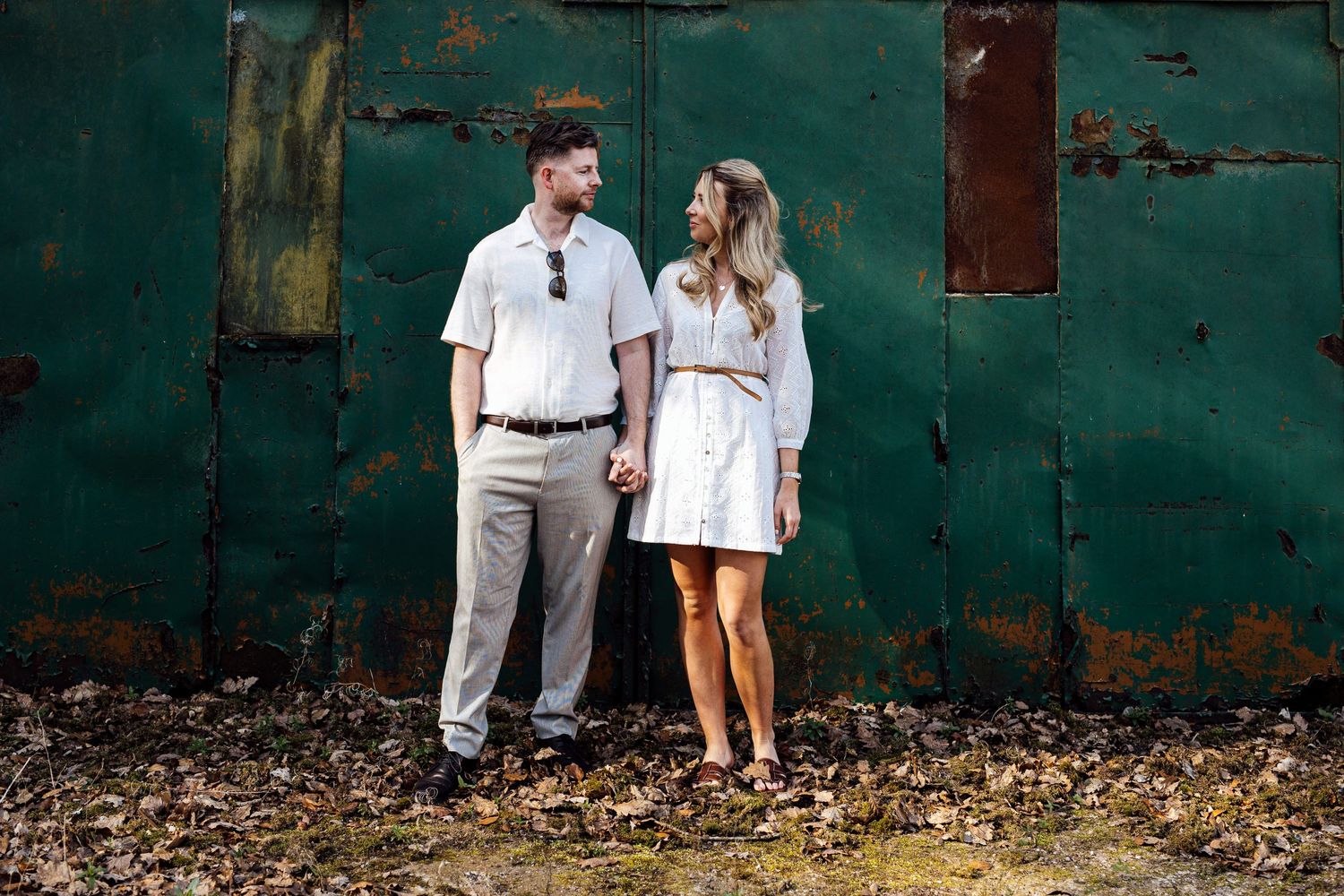A couple poses together against a weathered green wall during their engagement shoot.