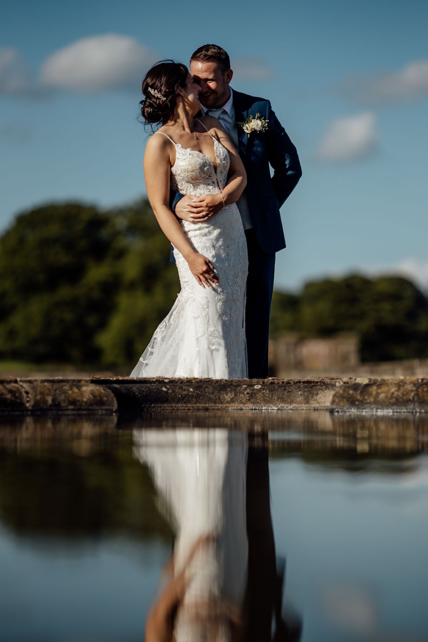 Couple in wedding attire photographed by reflective pond with blue sky backdrop.