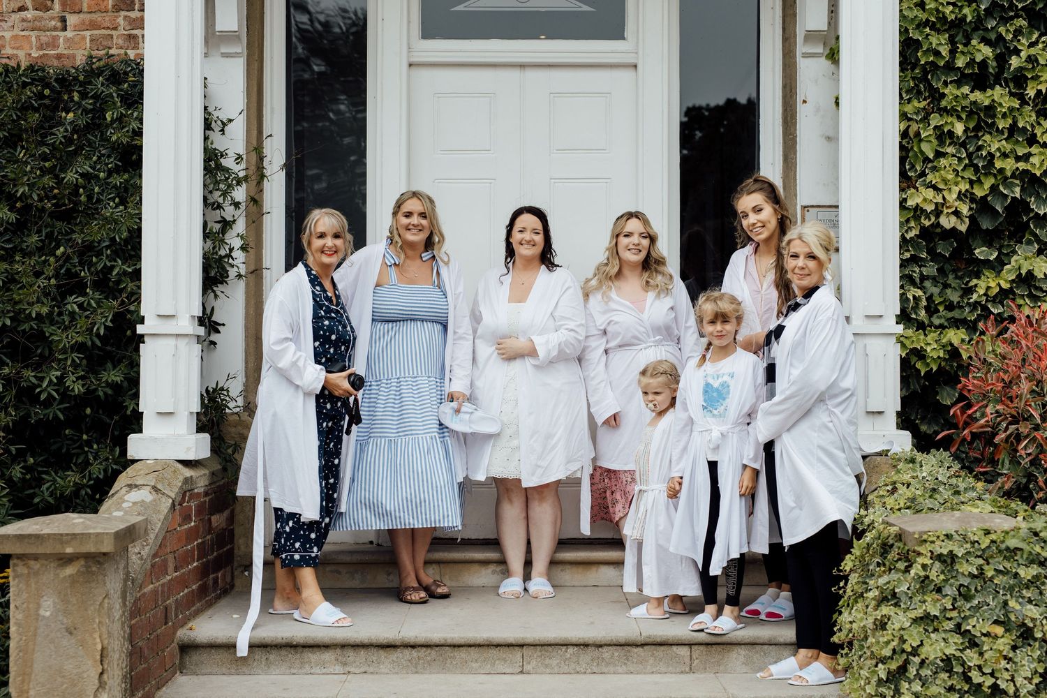 Wedding party in light blue and white dresses gathered on front porch steps of historic building.